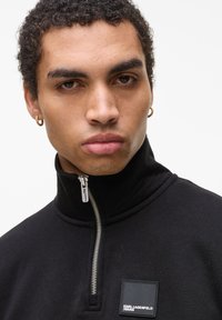 Felpa nera con zip e colletto alto, dotata di una zip argentata e di una toppa quadrata nera con testo bianco. I tratti del viso del modello sono visibili.