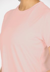 Vaaleanpunainen urheilupaita, joka on valmistettu pehmeästä ja hengittävästä kankaasta. Paidassa on lyhyet hihat ja pyöreä kaula-aukko. Edessä hienovarainen logoyksityiskohta.