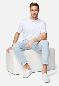 Jeune homme assis sur un bloc rectangulaire blanc, portant un t-shirt blanc, un jean bleu clair remonté et des baskets blanches.