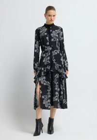 Schwarzes Kleid mit weißem Paisley-Muster, langen Ärmeln, gerafftem Saum und seitlichen Schlitzen. Kombiniert mit schwarzen Stiefeletten. Glatte Textur.