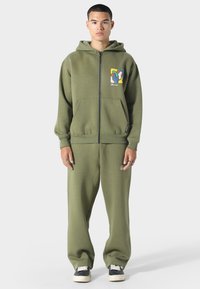 Olivengrøn zip-up hættetrøje med lomme foran og farverig abstrakt grafik; matchet med løstsiddende olivengrønne bukser og sorte sneakers.