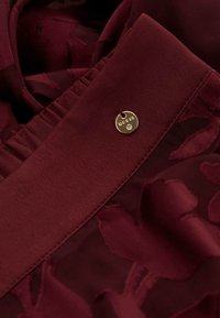 Bordo tkanina s cvjetnim uzorcima, sa širokim ovratnikom, nabranim detaljima i zlatnom oznakom s natpisom "GUESS" uz dijamantni naglasak.