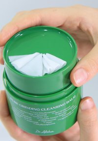 Zielony cylindryczny pojemnik na balsam oczyszczający z zakręcaną pokrywką, zawierający wewnątrz białą, teksturowaną maść. Etykieta z informacjami o produkcie na boku.