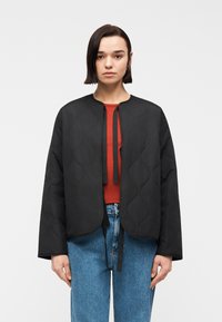 Νεαρή γυναίκα με ίσια σκούρα μαλλιά φοράει μαύρο καπιτονέ μπουφάν πάνω από κόκκινη μπλούζα και μπλε τζιν, στέκεται μπροστά σε λευκό, απλό φόντο.