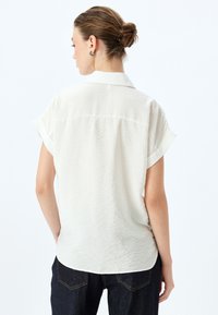 Camicia a maniche corte senza colletto in tessuto bianco sporco e strutturato. Lunghezza vita con maniche arrotolate, abbinata a jeans in denim scuro.