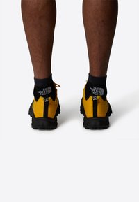 Gula sportskor med svarta detaljer och strukturerade sulor, som bärs med svarta ankelsockor med en logo. Visar en hållbar design.