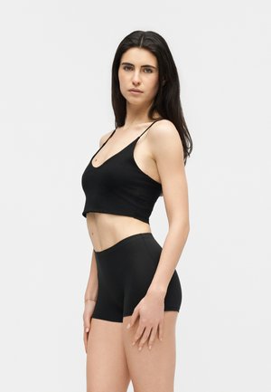 Donna con lunghi capelli scuri che indossa un top corto nero e pantaloncini neri a vita alta, in piedi davanti a uno sfondo chiaro e semplice.