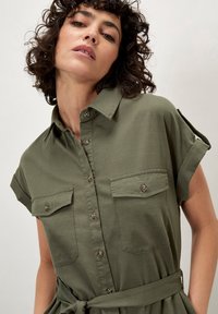 Olivenfarbendes Hemdkleid mit kurzen Ärmeln, durchgehender Knopfleiste, zwei Brusttaschen und einem gebundenen Taillendetail, aus einem leichten Stoff gefertigt.
