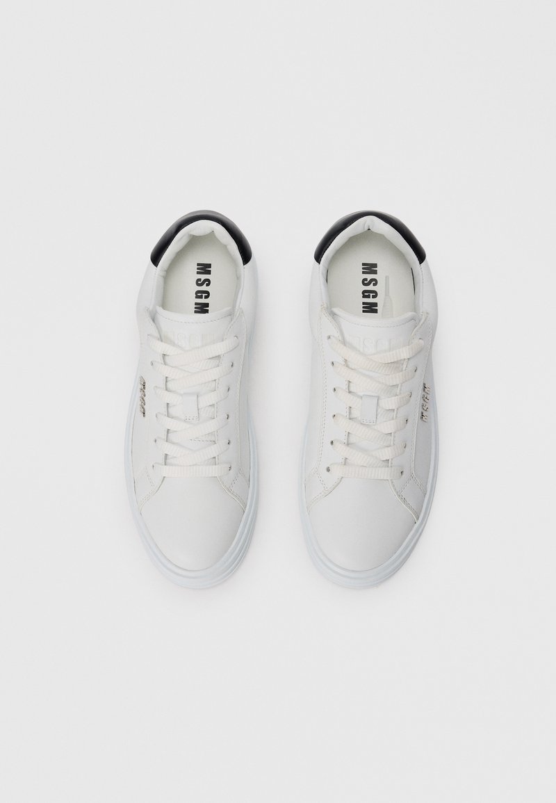 Une paire de baskets blanches basses avec des patchs noirs au talon, des lacets blancs et le logo "MSGM" sur les semelles beiges et sur le côté.
