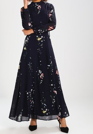 Femme portant une longue robe fleurie bleu marine à manches longues et des talons noirs pointus, debout devant un fond uni.