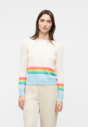 Kvinde med langt brunt hår iført en hvid sweater med horisontale regnbuestriber på bryst og ærmer, kombineret med lysebeige bukser.