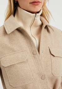 Béžová vlna směsová bunda s límcem, dvěma náprsními kapsami a zapínáním na zip u vysokého límce v žebrované pletenině.