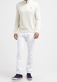 Vit stickad tröja med hög krage, dragkedjestängning, matchad med vita denimbyxor och marinblå sneakers med vita accenter.