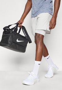 Μαύρη τσάντα duffel της Nike με γυαλιστερό λογότυπο, με διπλές λαβές και κύριο διαμέρισμα με φερμουάρ. Φορεμένη από άτομο με ανοιχτόχρωμο γκρι σορτς και λευκά αθλητικά παπούτσια.