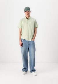 Camisa de botão de manga curta verde clara com um bolso no peito, combinada com umas calças de ganga azuis de corte largo e sapatilhas brancas. Boné verde na cabeça.