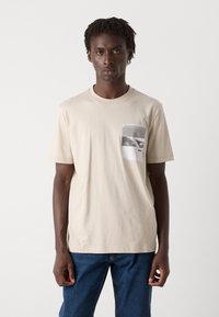 Beige bomulls t-skjorte med rund hals, korte ermer, og en frontlommepåtrykk med svart-hvite design. Brukt med blå jeans.