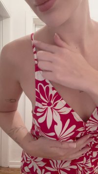 Czerwona sukienka w kwiaty z białymi wzorami liści, głęboki dekolt w kształcie litery V, cienkie ramiączka i dopasowany krój. W tle widać jasne wnętrze.