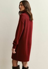 Robe en tricot à manches longues et col roulé de couleur rouge profond. Coupe ample avec des poignets et un ourlet en côtes. Texture douce, design jusqu'au genou.