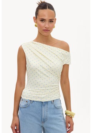 Femme aux cheveux foncés en chignon, portant des boucles d'oreilles créoles dorées, un top crème à une épaule avec des pois bleus, un jean bleu clair et des bracelets empilés.
