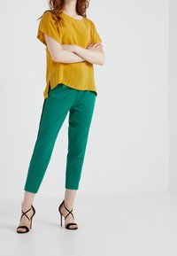Top giallo senape a vestibilità ampia con maniche corte, abbinato a pantaloni color teal con taglio affusolato, caratterizzati da strisce laterali nere, e tacchi neri con cinturini.