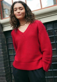 Ung kvinde med krøllet hår iført en rød ribbet V-hals sweater og sorte bukser, som står udendørs foran en murstensvæg og vinduer.