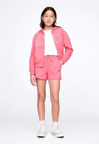 Ροζ φούτερ με το λογότυπο «GAP», συνδυασμένο με σορτς στο ίδιο χρώμα. Λευκό μπλουζάκι από μέσα. Το μοντέλο φοράει λευκά αθλητικά παπούτσια υψηλού άξονα.