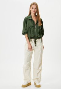 Groene cropped jas met borstzakken en zilveren hardware, gecombineerd met losse beige broek en chunky crèmekleurige sneakers.