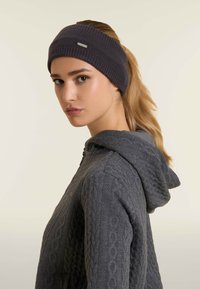 Bandeau en tricot gris avec un motif texturé, arborant un subtil logo métallique. Porté avec une veste à capuche grise.