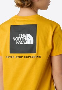 Camiseta de algodón amarilla con un gráfico rectangular negro que muestra el logo de "THE NORTH FACE" y el texto "NEVER STOP EXPLORING" en la parte trasera.