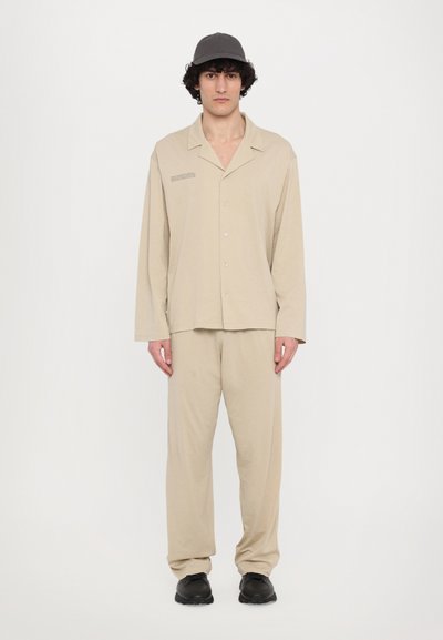 Beige Baumwollhemd mit langen Ärmeln und lässigem Schnitt, kombiniert mit passenden beigen Hosen. Schwarze Schuhe und eine graue Mütze runden den Look ab.
