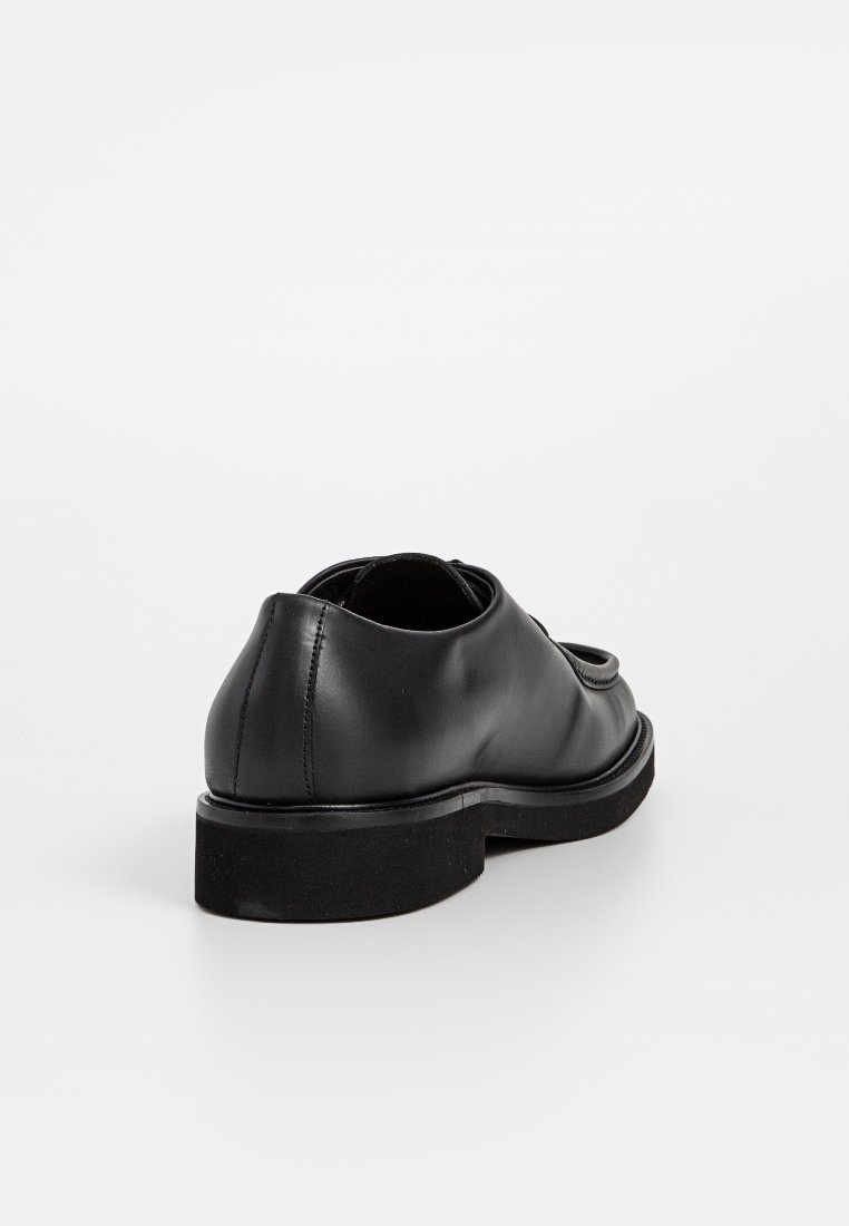 Zapato de cuero negro con una textura lisa, punta redondeada y suela baja y gruesa. Diseño minimalista sin hardware ni adornos visibles.
