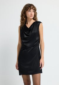 Vestido de satén negro con un escote en cowl drapeado, diseño sin mangas y textura suave; el dobladillo cae a la mitad del muslo. Silueta simple y elegante.