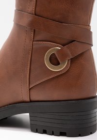 Bottine en cuir marron avec une sangle et un détail en anneau en laiton, présentant un petit talon noir empilé avec un semelle striée pour une meilleure adhérence.