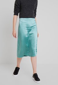 En satin, knälång kjol i en livlig turkos färg, med en slät yta och en lätt kurvad fåll, parat med svarta sneakers.
