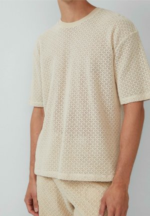 Homme portant un haut beige à manches courtes en maille filet et un short assorti sur un fond clair uni.