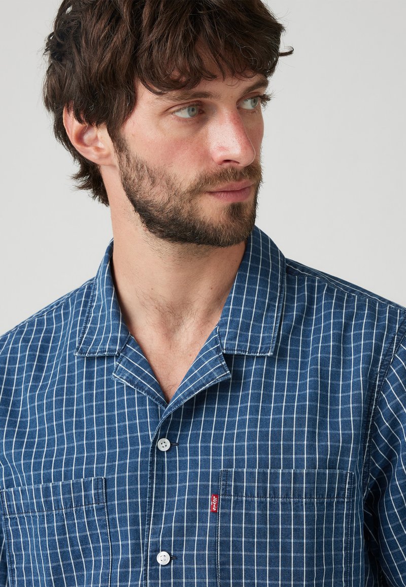 Camisa de manga corta hecha de algodón azul con un patrón de cuadrícula blanca. Presenta un cuello tipo campamento, botones en la parte delantera y un bolsillo en el pecho izquierdo con un logo.