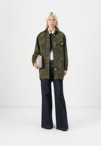 Olivgrüne Cordjacke mit zwei Brusttaschen, kombiniert mit einem weißen Hemd und einer schwarzen Krawatte, weit geschnittenen dunkelblauen Jeans, hält eine lavendelfarbene Tasche.