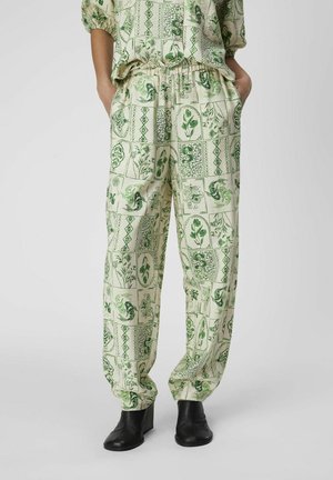 Pantalon ample crème avec imprimé botanique et géométrique vert, porté avec des bottines noires et les mains dans les poches.
