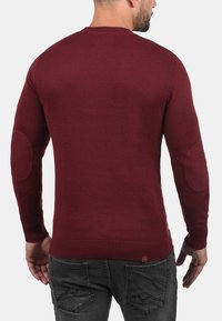 Bordeauxfarbener Strickpullover mit gerippten Bündchen und Saum. Verfügt über Ellbogenflicken und einen klassischen Rundhalsausschnitt. Glatte Textur mit figurnahem Schnitt.
