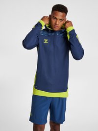 Marinblå och limegrön huvtröja i sportig stil med prickigt mönster, långa ärmar och logotyp; matchad med marinblå shorts. Mjuk textur.