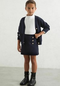 Námornícky cardigan s akcentmi zlatých gombíkov cez biely rolák, skombinovaný s námorníckou mini sukňou so zlatými gombíkovými detailmi a čiernymi členkovými čižmami.