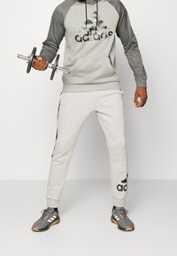 Sweat à capuche gris et gris foncé avec un grand logo noir, associé à un pantalon de survêtement gris clair avec une bande noire et un logo. Porte des baskets grises.