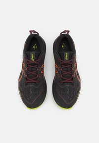 Svarte treningssko med teksturert stoff, lyse oransje detaljer, bordeauxsnører og gulgrønne såler. Designet for ytelse og støtte.