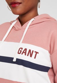 Sweatshirt à capuche rose avec des rayures horizontales blanches et marine. Comprend le logo GANT en bordeaux, des cordons de serrage et une texture de tissu douce.