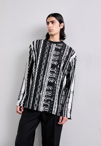 Čierno-biely dlhý rukáv s odvážnymi grafickými vzormi, vertikálnymi pruhmi a textom, s voľným strihom a okrúhlym výstrihom.