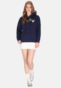 Sudadera con capucha azul marino con cordones y texto "Dino Club" en rosa, con detalles de flores. Combinada con una minifalda blanca y zapatillas grises.