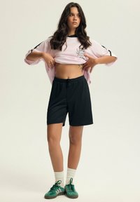 Różowa, oversize'owa koszulka sportowa z graficznym nadrukiem, połączona z czarnymi szortami. Model nosi zielone buty i białe skarpetki. Gładka faktura na całej powierzchni.