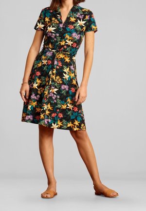 Kvinde iført en kortærmet, knælængde sort kjole med flerfarvet tropisk blomsterprint og et bindebånd i taljen, kombineret med brune slidesandaler.