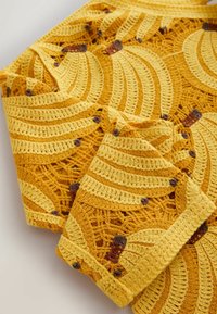 Hæklet stof med gule og gyldne bananmønstre med brune stilke på en tekstureret åbenvævet baggrund.