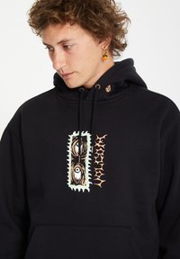 Zwarte hoodie gemaakt van zacht materiaal met een vooraanstaand grafisch ontwerp in wit, groen en oranje, voorzien van abstracte vormen en oogmotieven.